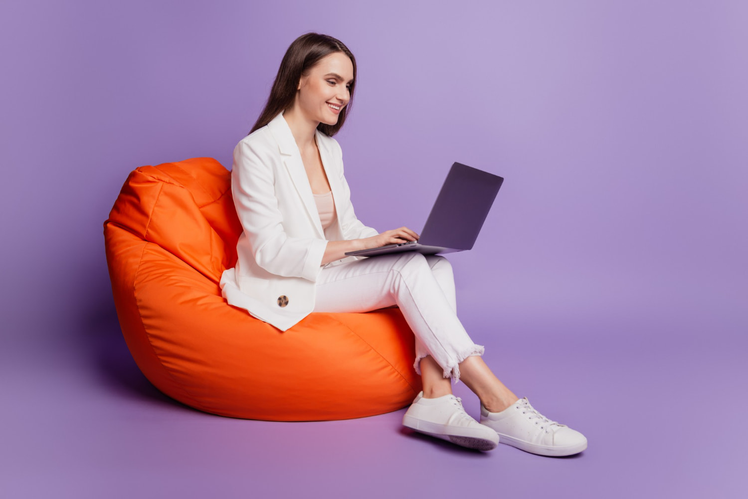 Mulher branca de cabelo médio castanho, sentada em um puf laranja com um laptop na perna.