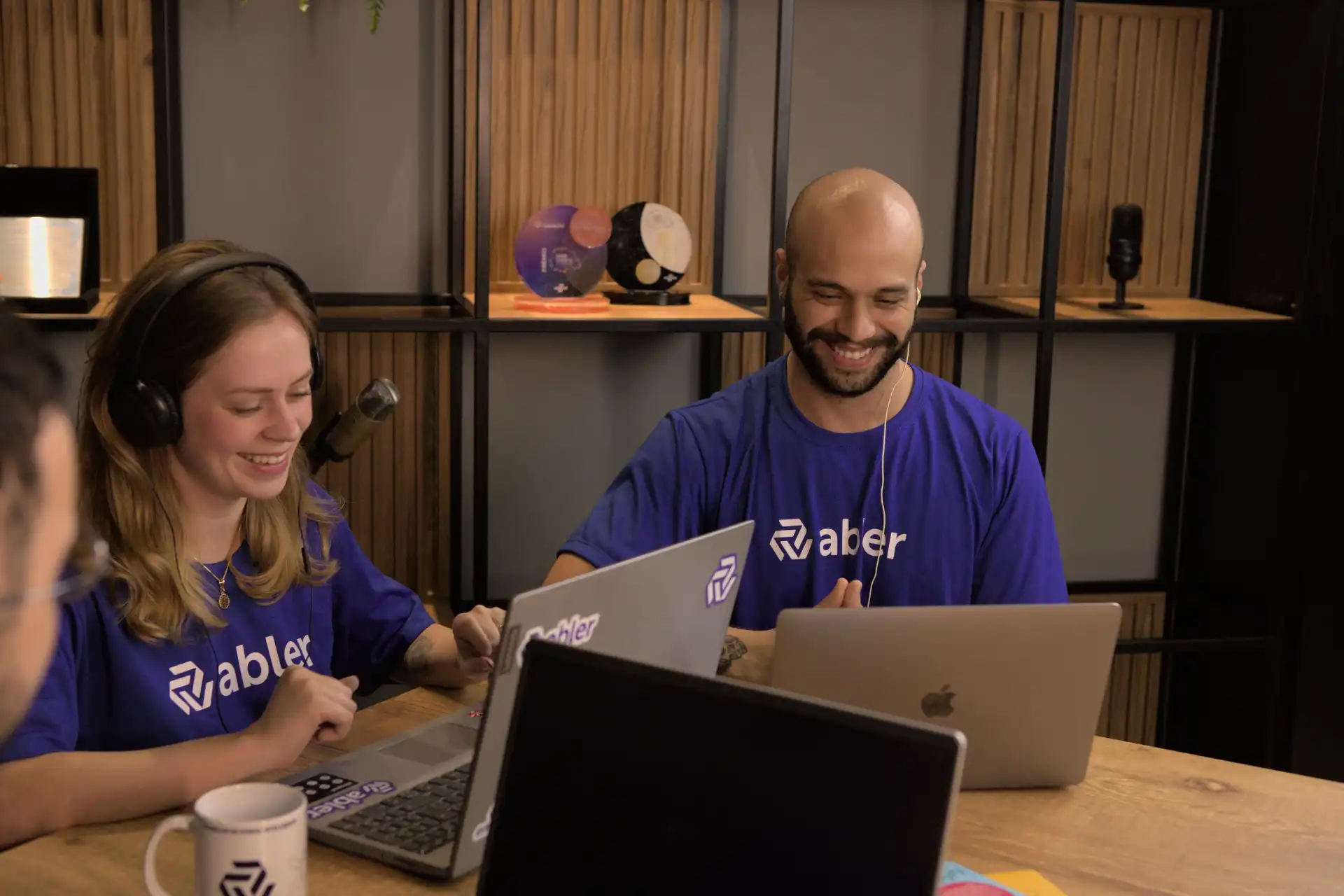 Imagem de profissionais sorrindo em um ambiente de trabalho colaborativo, realizando recrutamento online. A equipe está usando laptops e fones de ouvido, enfatizando a tecnologia no processo de seleção.