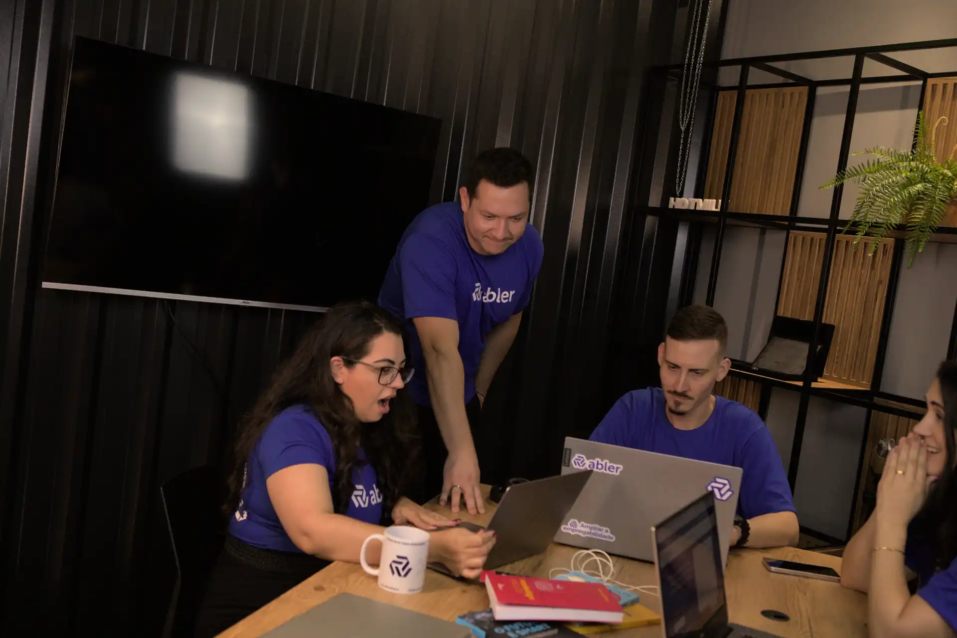 Grupo de profissionais discutindo desafios no recrutamento e seleção em um ambiente de trabalho moderno, com laptops e uma tela ao fundo.