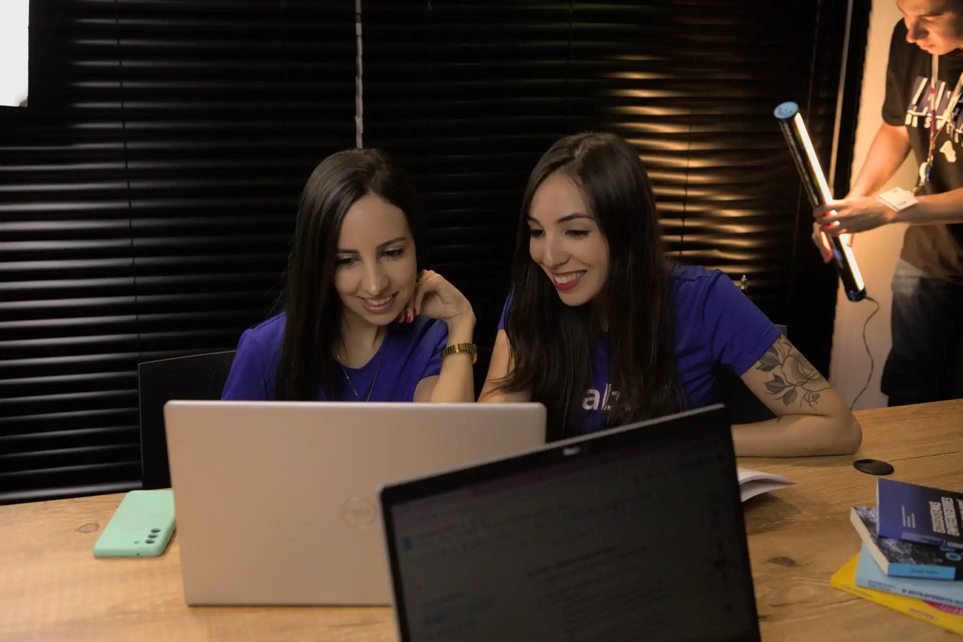 Duas mulheres sorrindo enquanto participam de um processo seletivo interno, analisando conteúdo em um laptop e se envolvendo em atividades colaborativas.