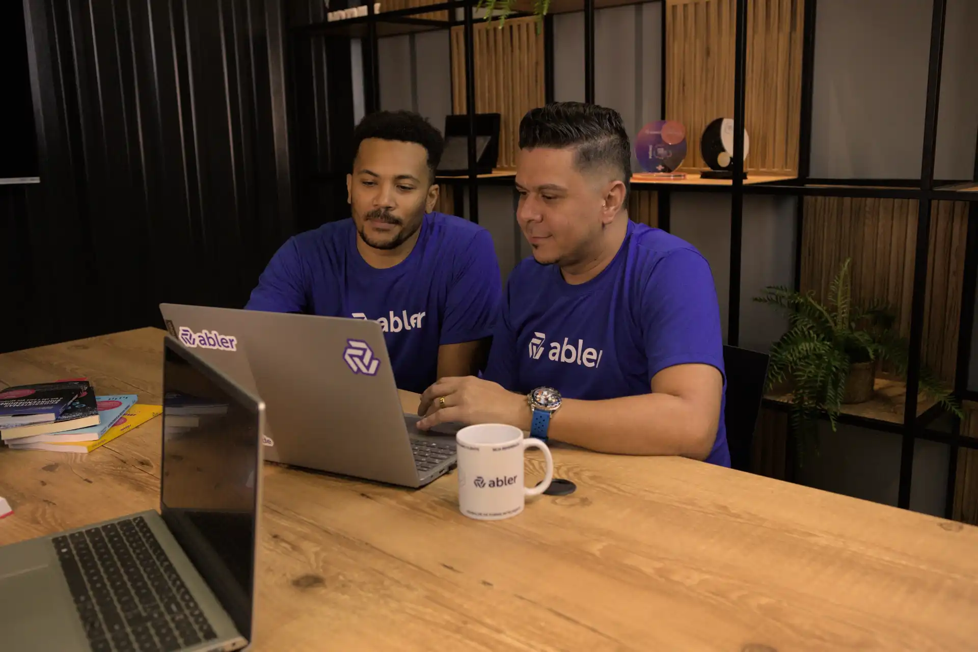 Dois homens em camiseta azul da empresa abler, analisando informações de parecer de candidato em um laptop durante uma reunião de trabalho em um ambiente moderno.