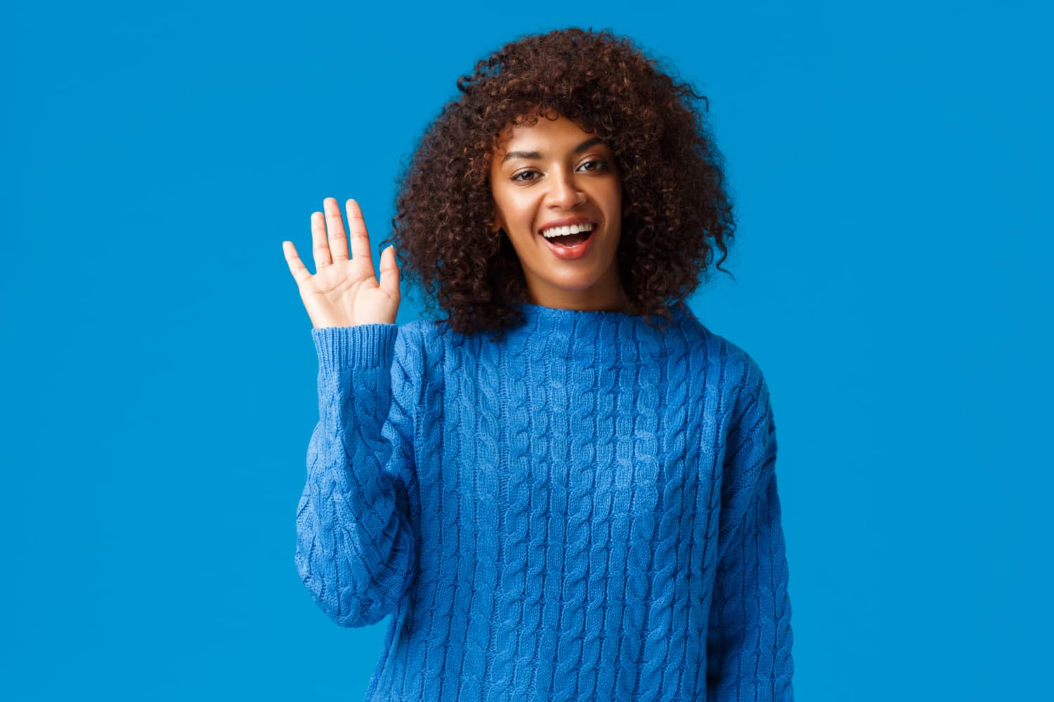 mulher negra de cabelo blackpower dando tchau e sorrindo, usando uma camisa azul em um fundo azul.