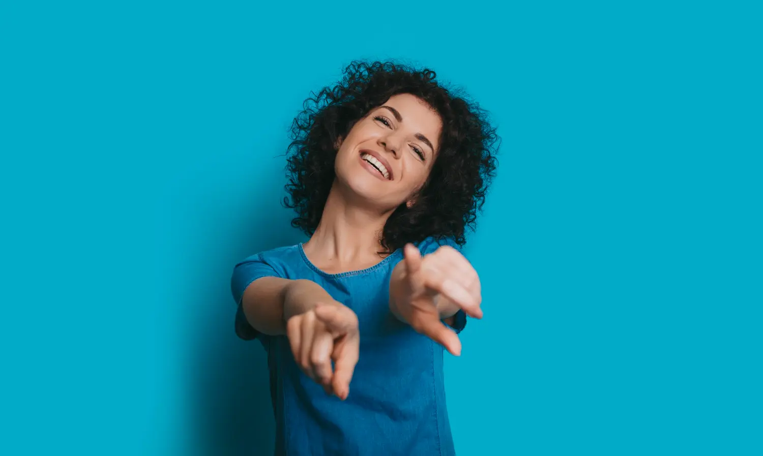 mulher branca de cabelos pretos encaracolados curto, camiseta azul, em um fundo azul, apontando para você.