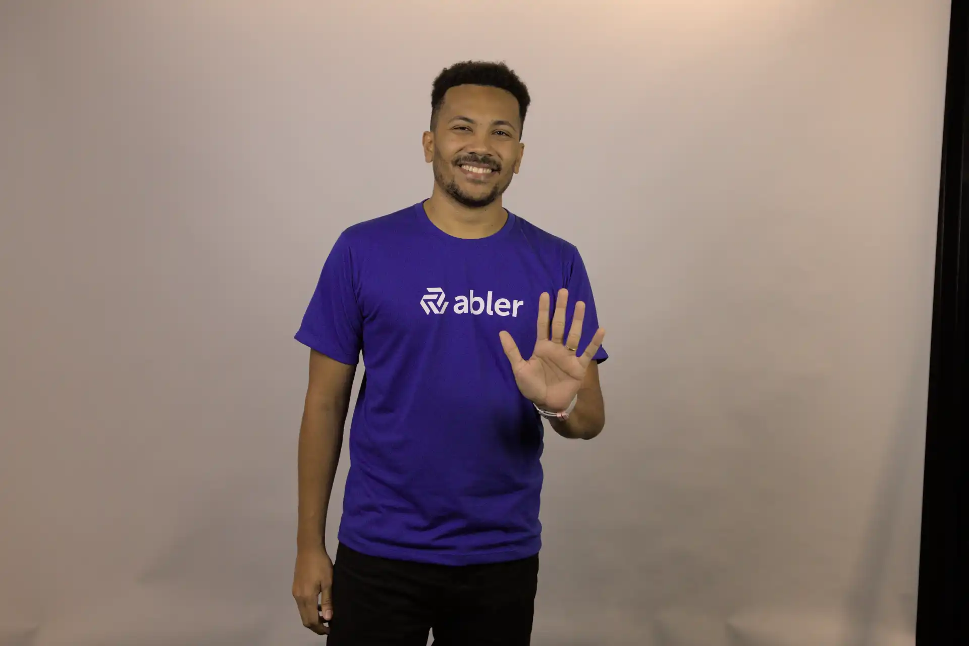 Homem sorridente vestindo camiseta roxa da marca Abler, fazendo sinal de adeus em um fundo neutro. Imagem promovendo resoluções pessoais e profissionais.