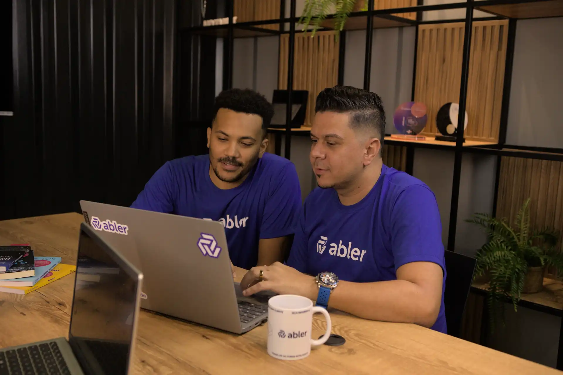 Dois homens analisando perfil comportamental em um laptop em uma sala moderna, com materiais de apoio como livros e canecas sobre a mesa.