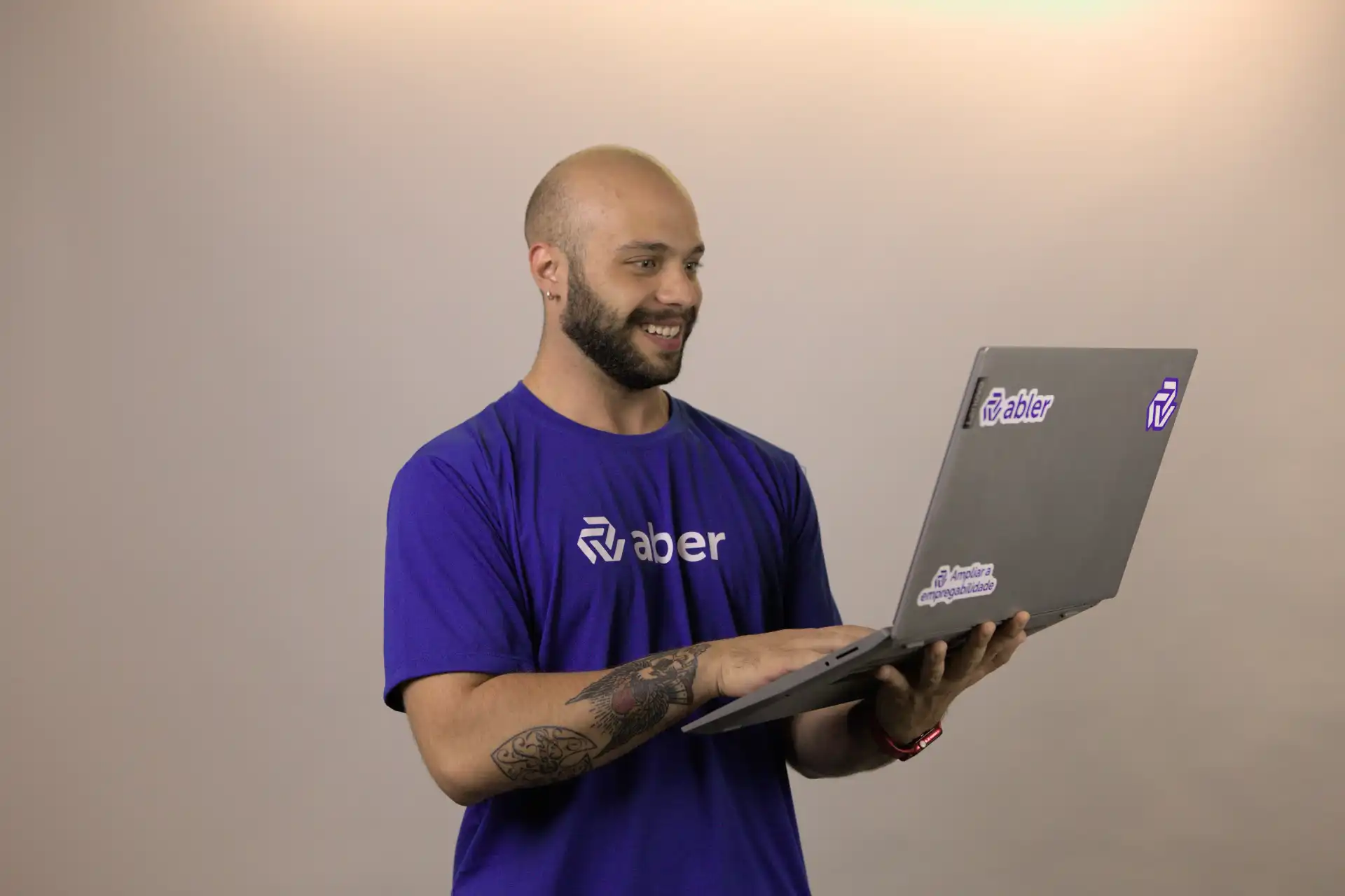 Homem sorridente com tatuagens, usando camiseta azul da empresa Abler, trabalhando em um laptop. A imagem representa desafios no recrutamento.