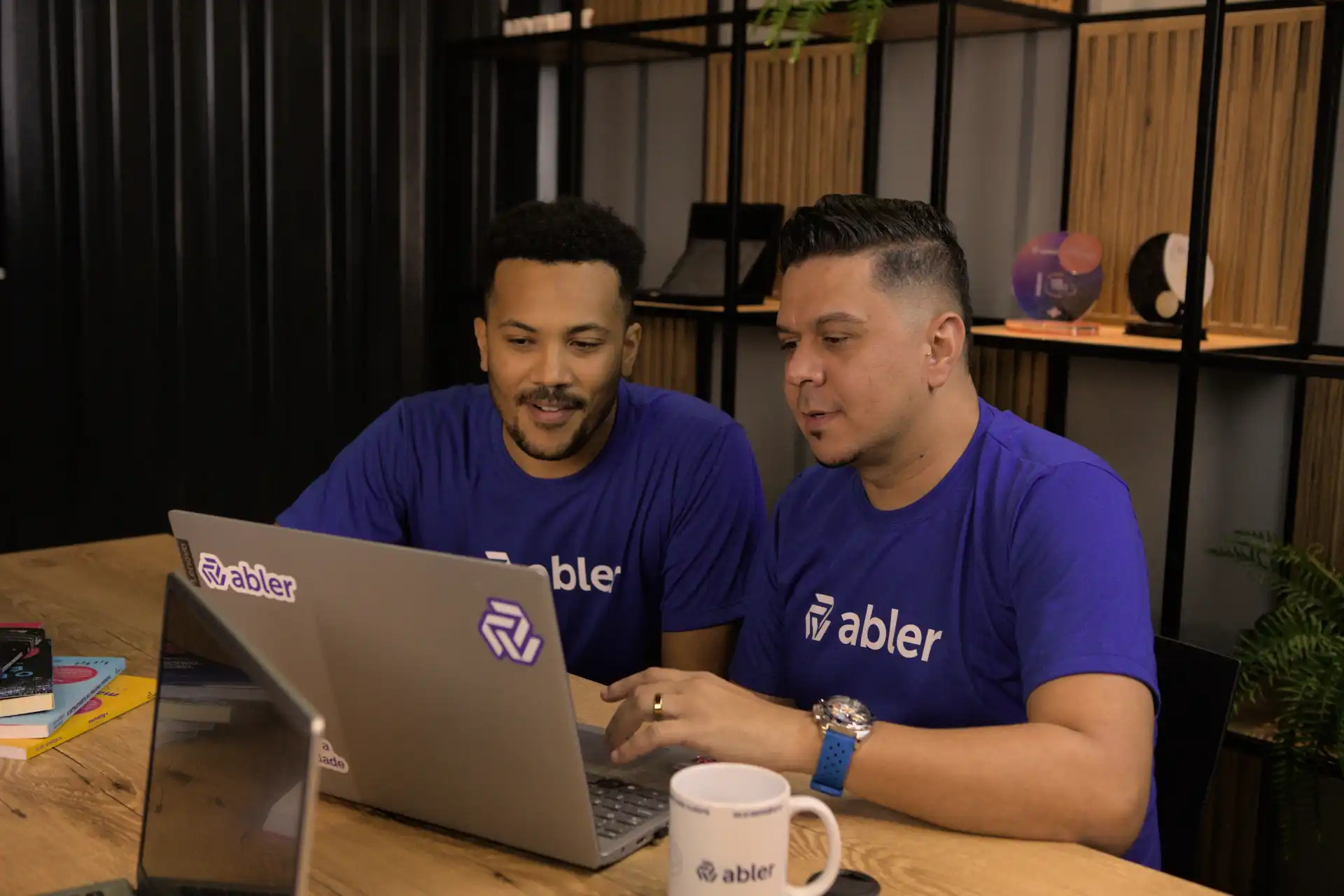 Dois homens com camisas azuis da abler analisando informações sobre regime CLT em um laptop durante uma reunião produtiva em um ambiente moderno.