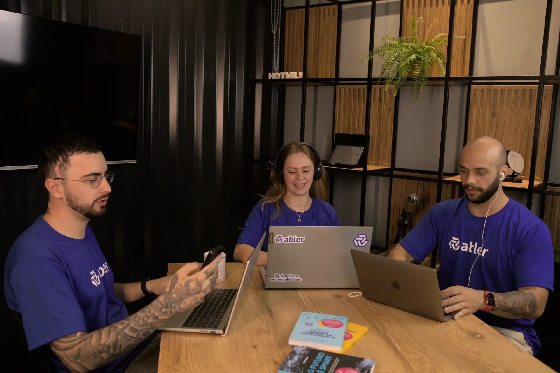 Equipe trabalhando em um ambiente colaborativo, focando em estratégia de recrutamento por indicação. Três profissionais com laptops e anotações em mesa.