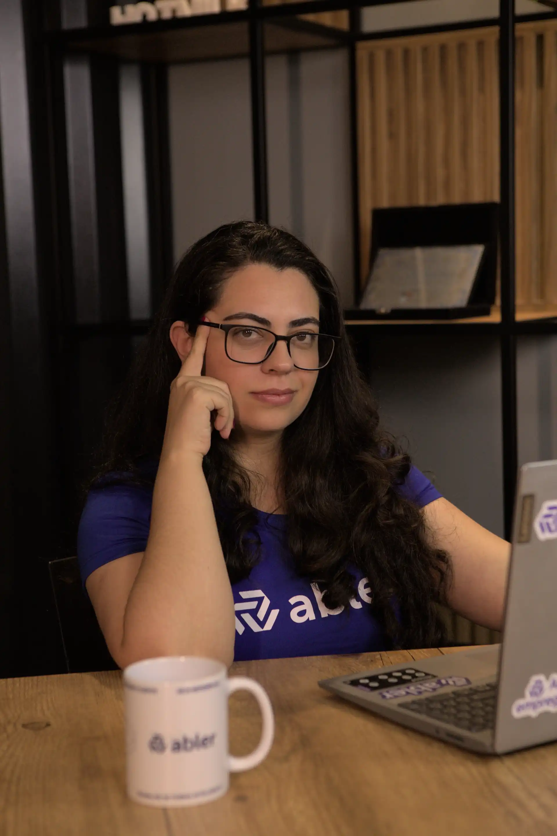 Mulher com camiseta azul da empresa abler, pensativa, sentada em uma mesa de trabalho com um laptop e caneca, representando os desafios dos profissionais autônomos.