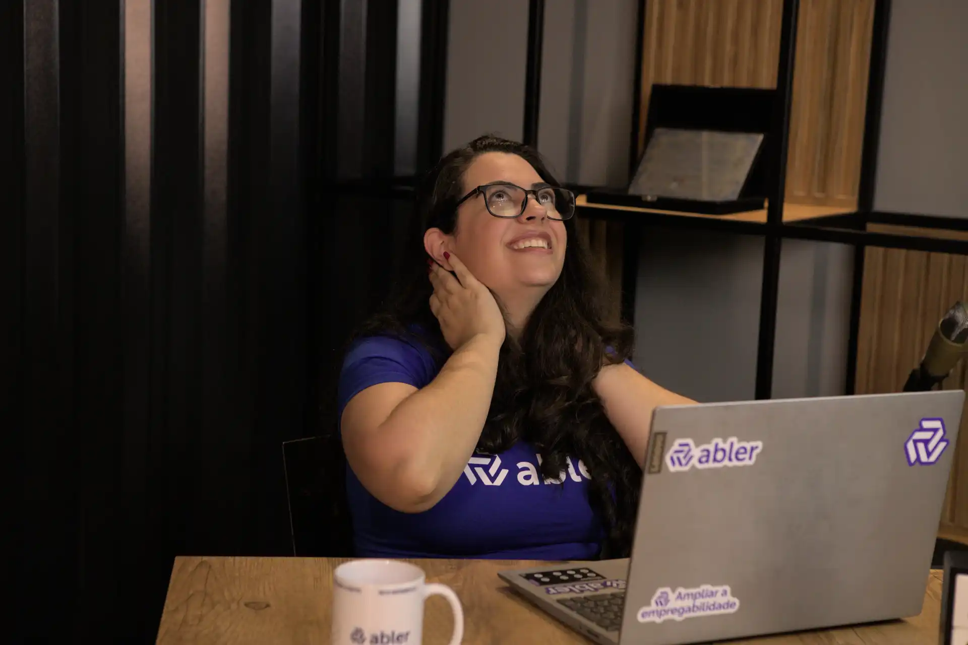 Imagem de uma mulher sorridente, com óculos e camiseta da Abler, refletindo sobre o processo de recrutamento e seleção enquanto trabalha em seu computador.