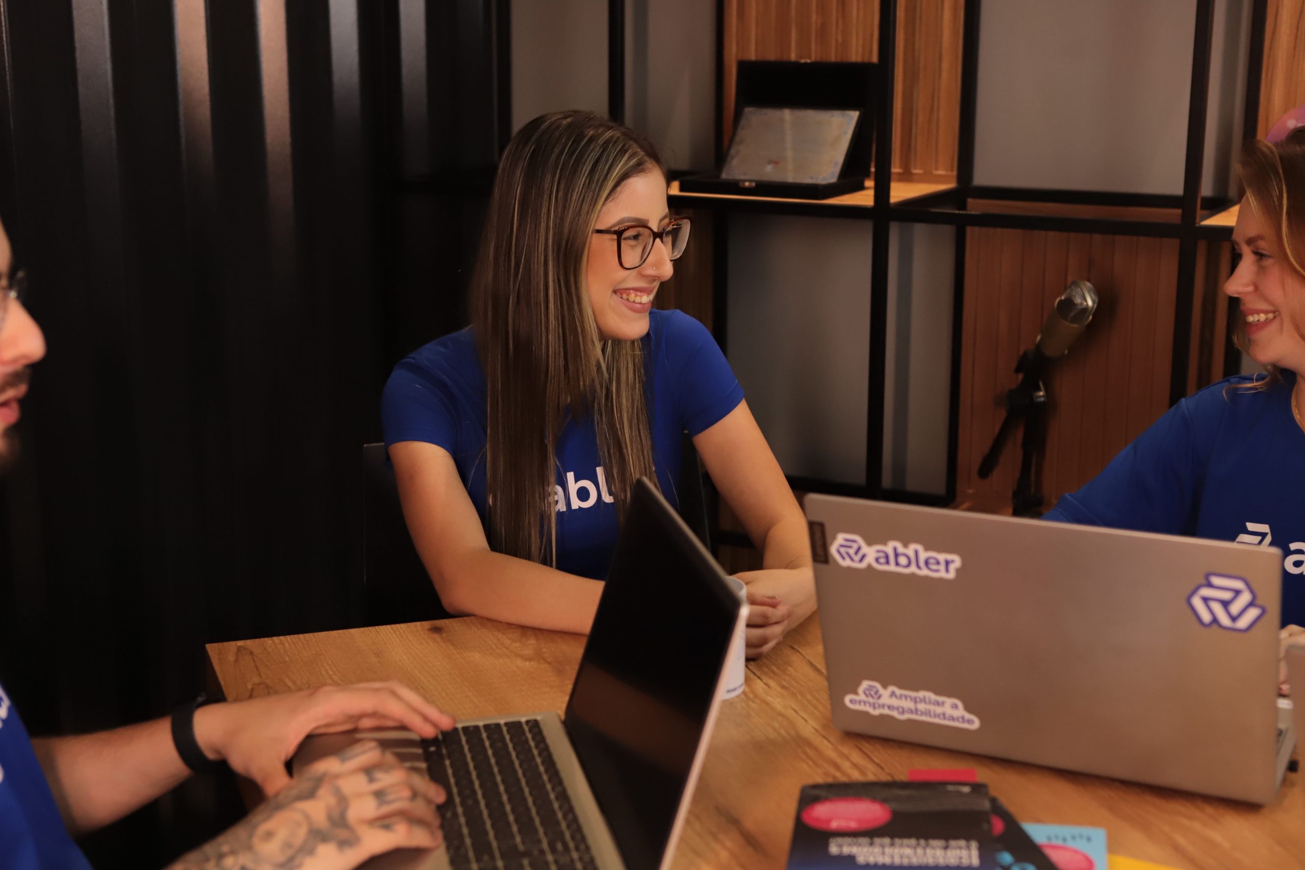 Três pessoas em uma mesa de madeira em um escritório com iluminação quente; duas mulheres ao centro e à direita conversam e sorriem, usando camisetas azuis da abler, enquanto trabalham em laptops com adesivos da marca. À esquerda, parte de um homem digitando em outro notebook. Ao fundo, há uma estante com divisórias, um microfone e um laptop aberto, sugerindo um ambiente de trabalho colaborativo e de produção de conteúdo.