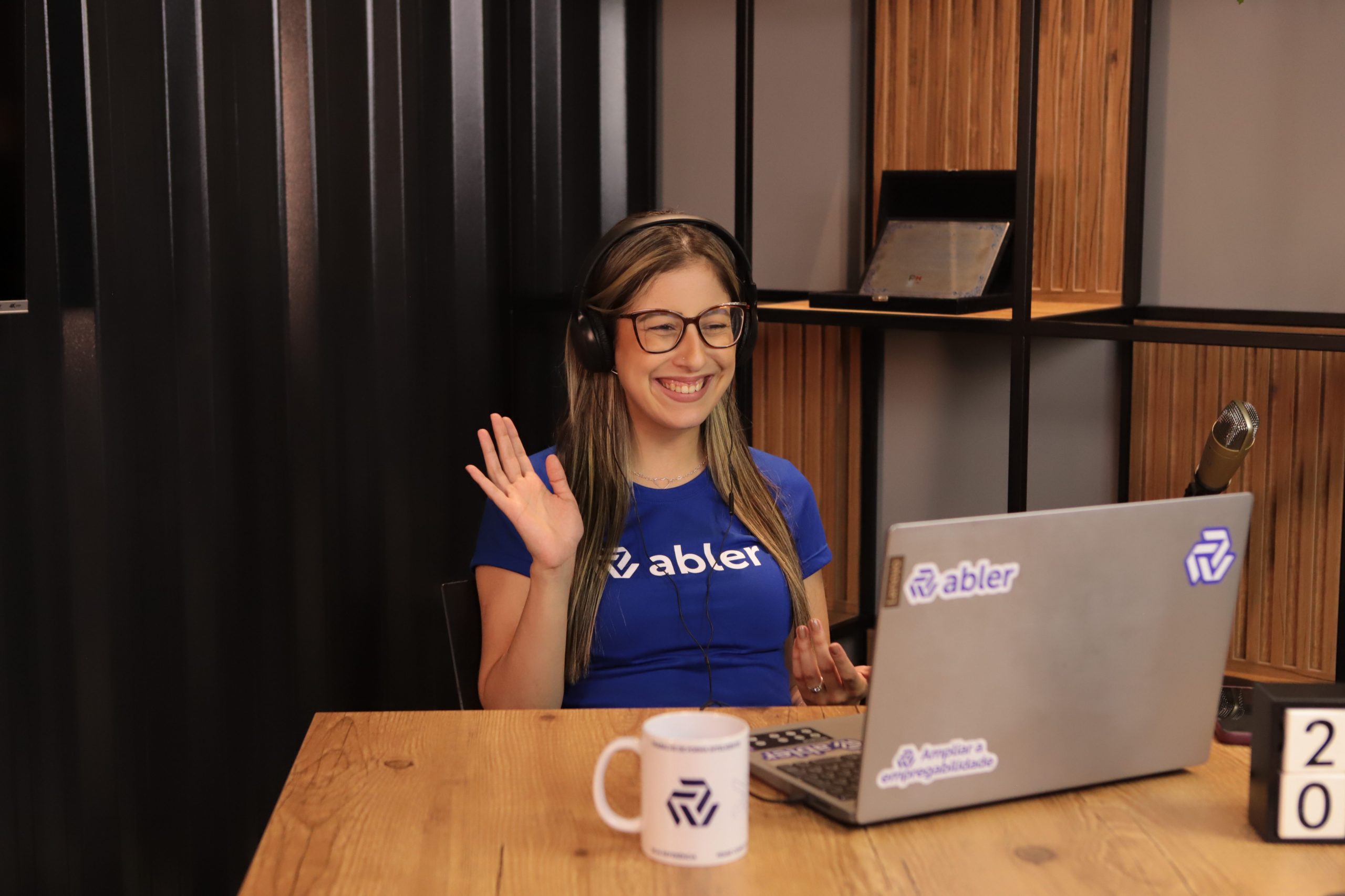 Mulher sorrindo em uma mesa de madeira, usando fones de ouvido e acenando com a mão durante uma videochamada. À sua frente há um notebook com adesivos da abler e uma caneca sobre a mesa. Ao fundo, há uma estante com detalhes em madeira e uma parede com cortina escura.