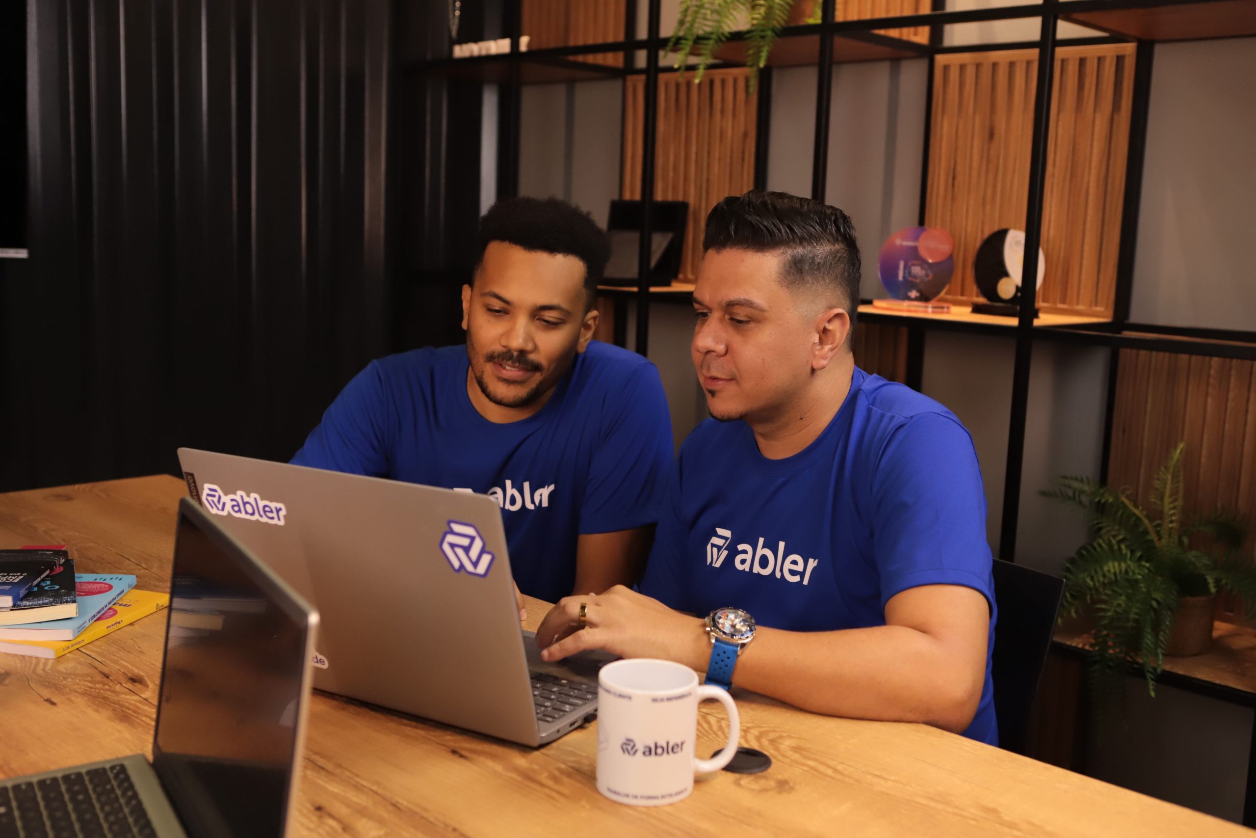 Duas pessoas usando camisetas azuis com o logotipo da abler trabalham juntas em um notebook sobre uma mesa de madeira, analisando perfis e realizando o sourcing de candidatos de forma colaborativa. Ao lado, há uma caneca com a marca e outro notebook parcialmente visível, enquanto ao fundo uma estante moderna com plantas e objetos decorativos compõe o ambiente. A cena representa o processo de sourcing como parte de uma infraestrutura de recrutamento previsível, saindo do modelo passivo de espera para uma abordagem ativa e estratégica de busca de talentos.