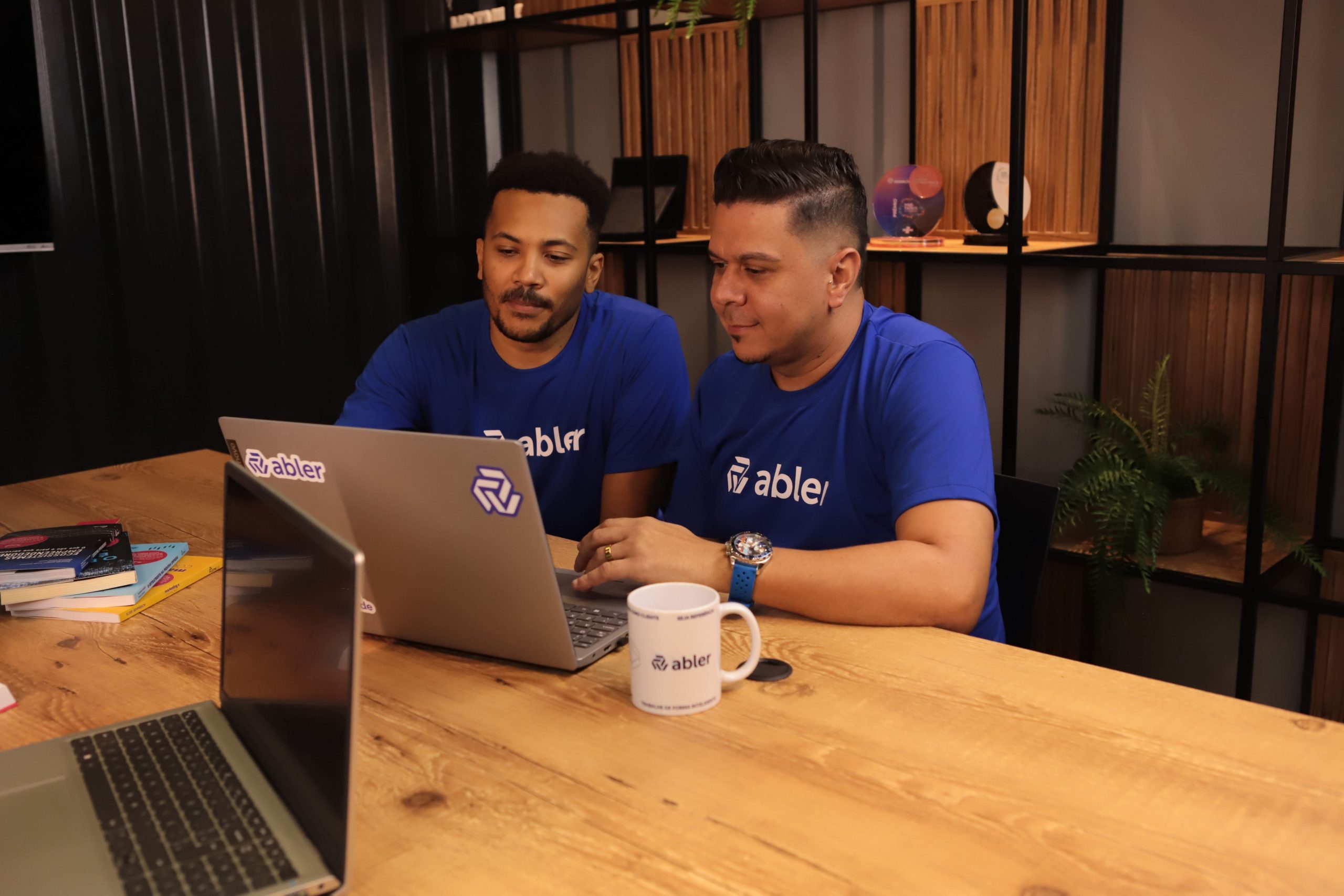 Dois profissionais usando camisetas azuis da abler trabalham juntos em frente a um notebook sobre uma mesa de madeira, em um ambiente de escritório moderno. Um deles aponta para a tela enquanto o outro observa com atenção, sugerindo colaboração e análise de dados. Ao fundo, há uma estante com objetos decorativos e plantas. A cena representa um momento de alinhamento e estratégia relacionado à construção de um funil de talentos.