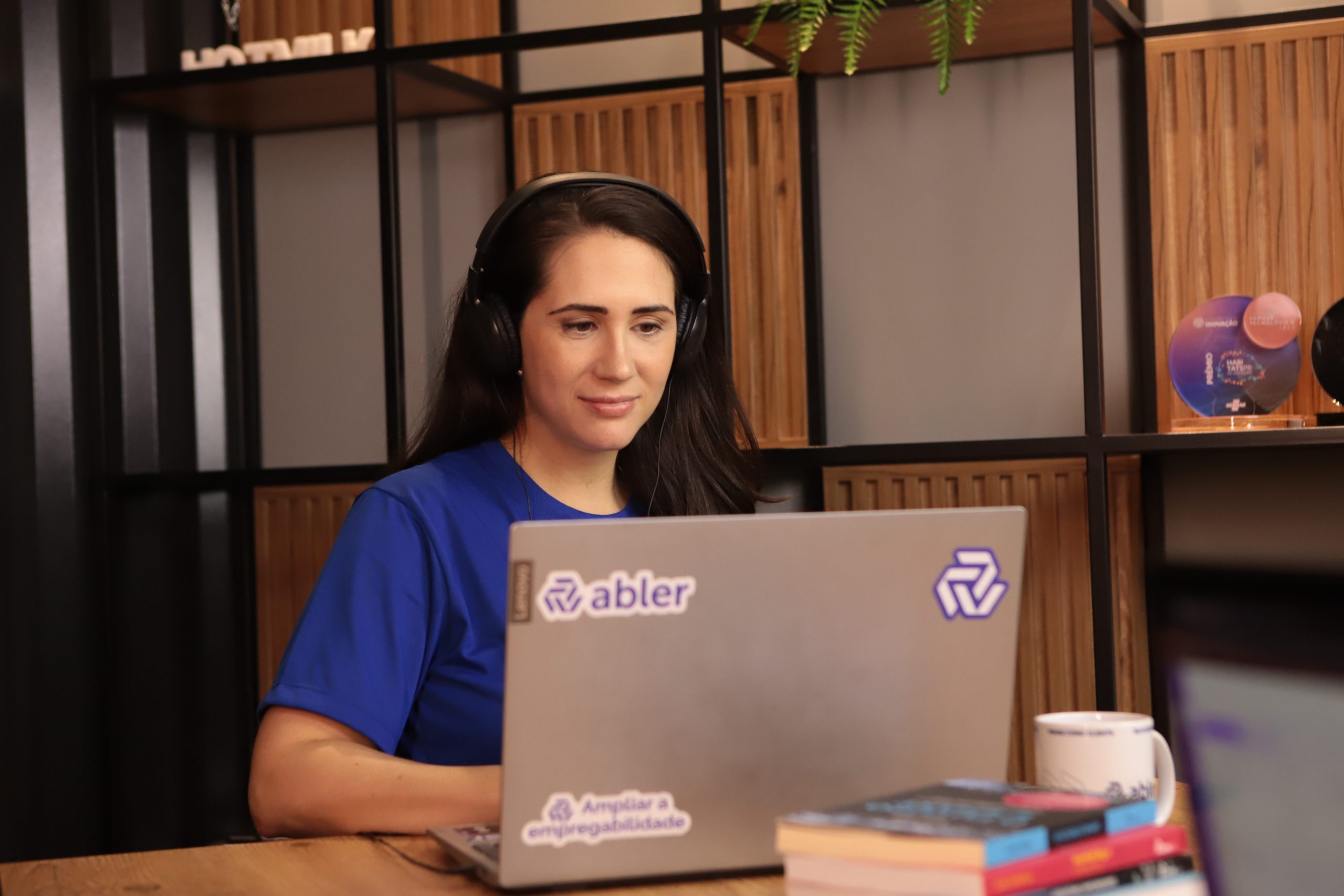 Mulher usando fones de ouvido trabalha em um laptop com adesivo da abler, sentada em uma mesa de madeira em um ambiente de escritório moderno, com estantes ao fundo e alguns livros e uma caneca sobre a mesa.