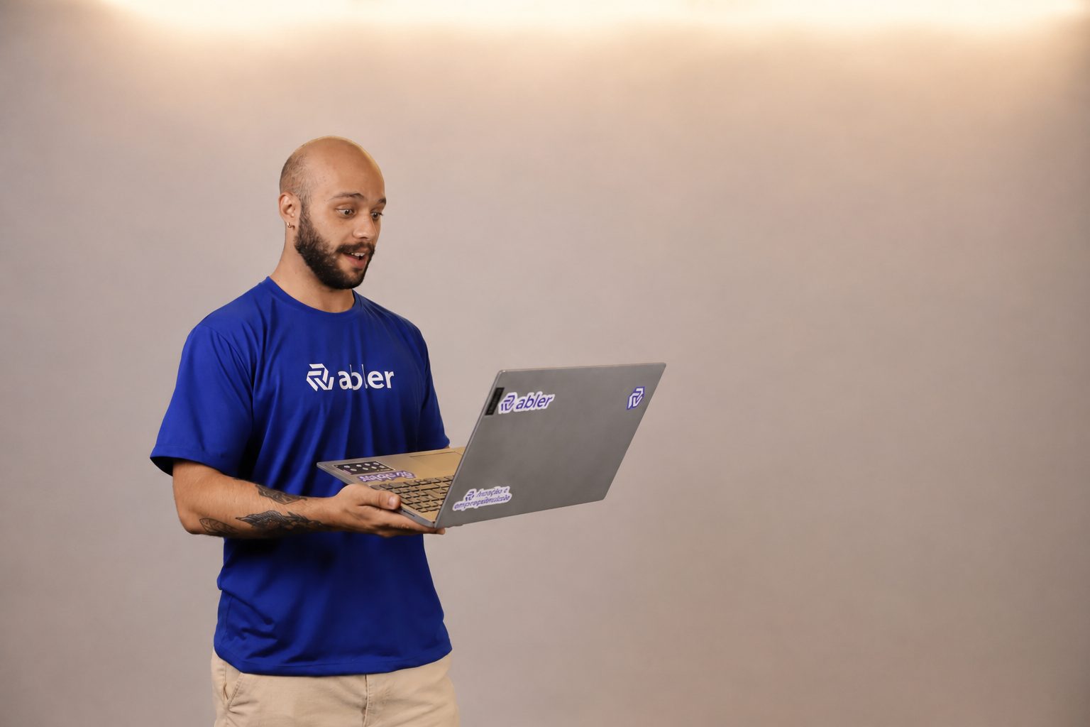 Profissional vestindo camiseta azul da abler segura um notebook com expressão de surpresa positiva enquanto observa a tela, em fundo neutro e amplo, representando o impacto da tecnologia no recrutamento, automação de processos seletivos, inovação em RH e os desafios entre eficiência operacional e caos digital na gestão de talentos.